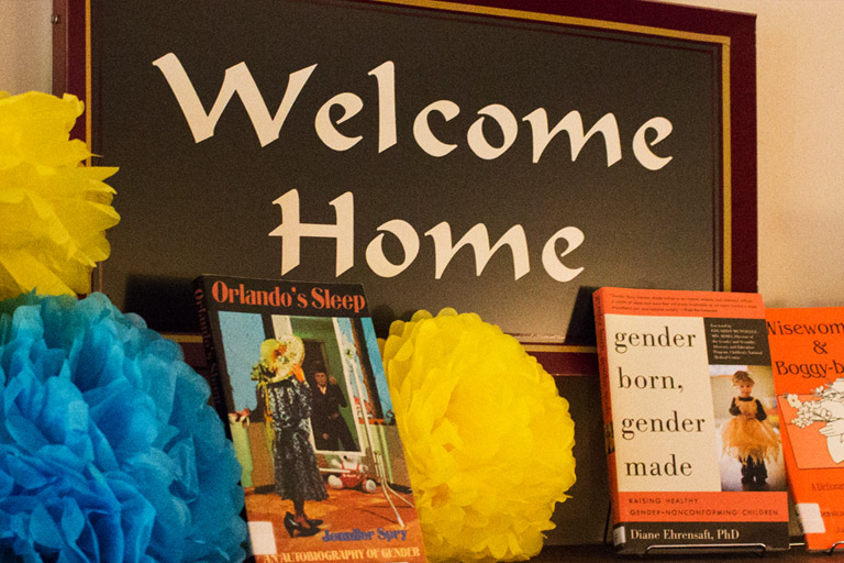 A sign in the LGBTQ+ Culture Center that says "Welcome Home". It is surrounded by LGBTQ+ books and other decorations.